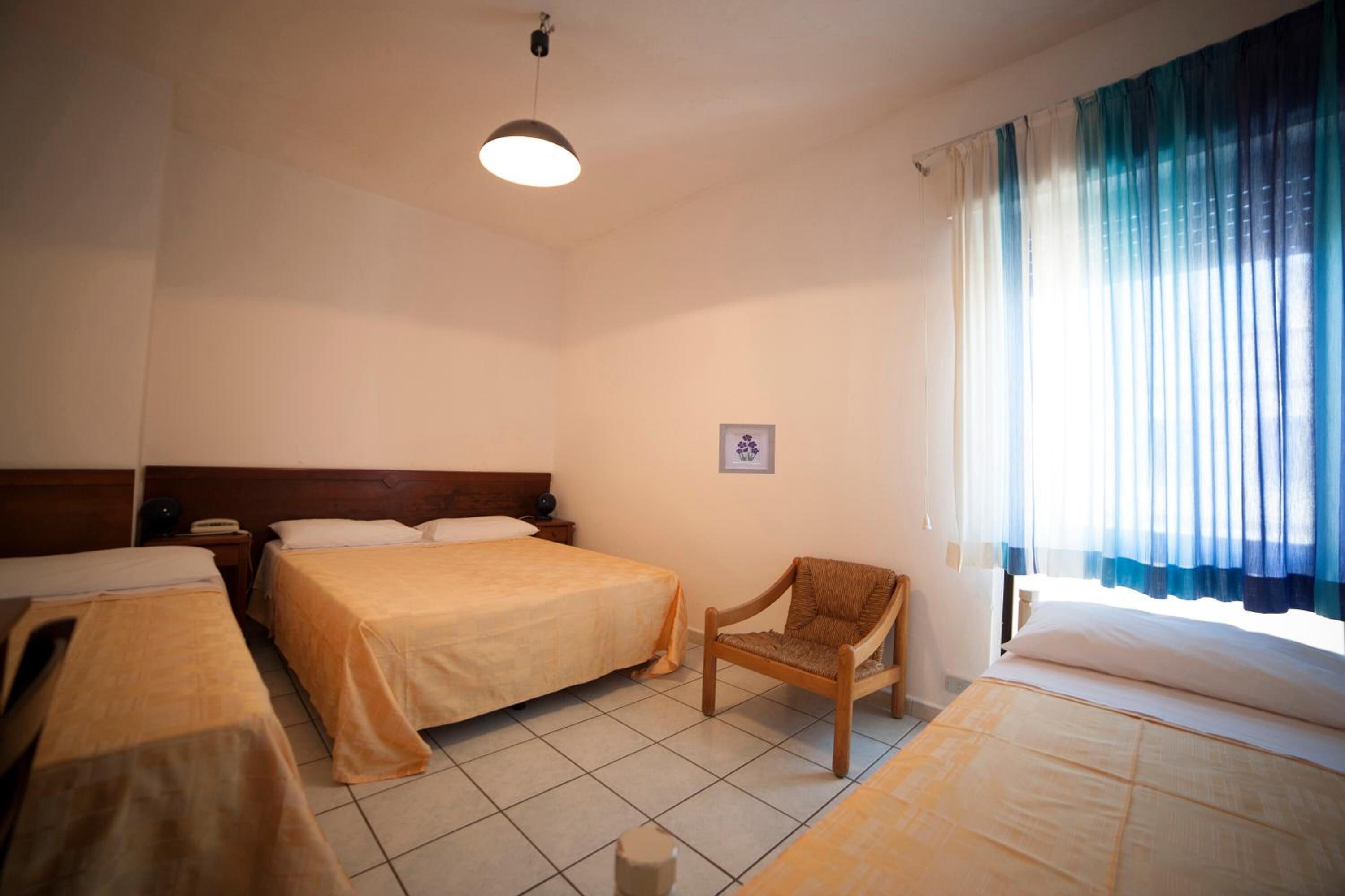 Une chambre d’hôtel avec trois lits, une chaise en bois, un luminaire au plafond et une fenêtre avec des rideaux bleus.