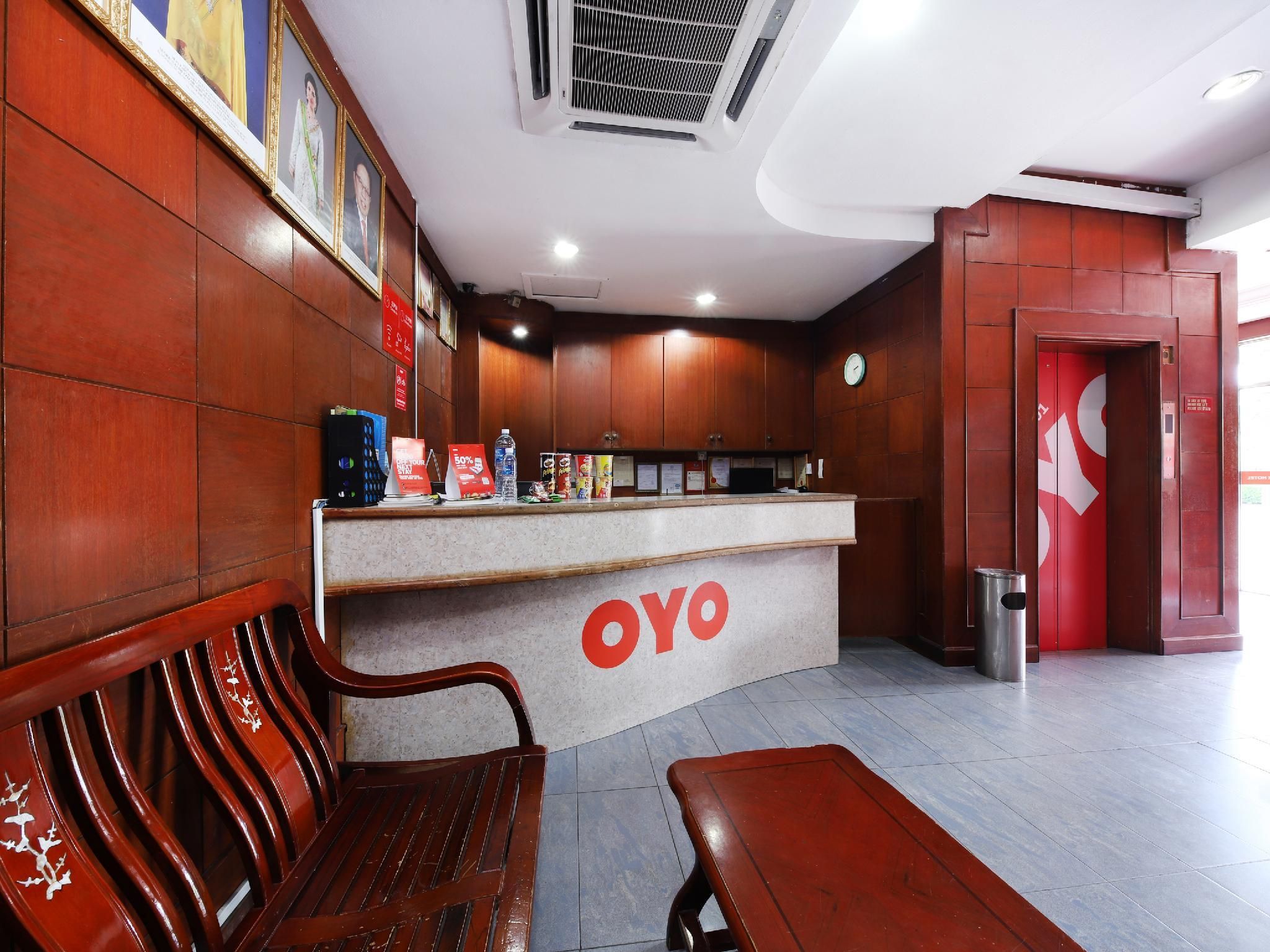 A hotel lobby with a reception desk labeled 'OYO,' wooden benches, a trash bin, and a clock on the wall.