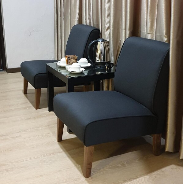 Ein modernes Zimmer mit einem dunkelblauen Sofa und passendem Sessel, einem kleinen Tisch mit Wasserkocher und Tassen sowie einem Vorhang im Hintergrund.