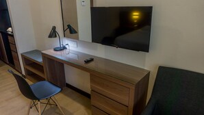 Una habitación compacta con escritorio de madera, una silla, un televisor montado en la pared y un estante pequeño.