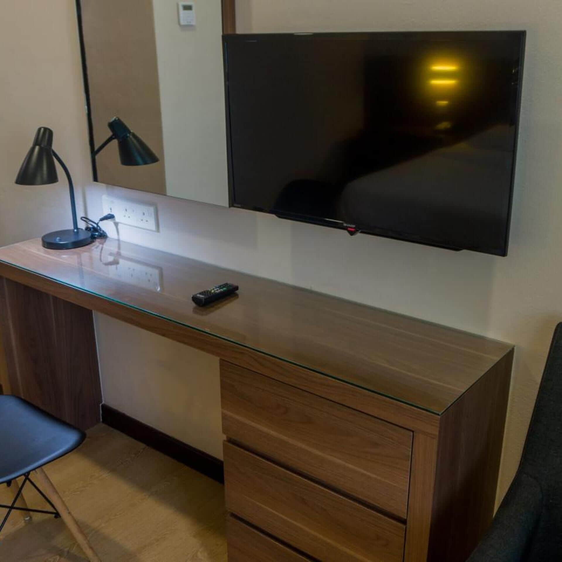 A modern office space with a wooden desk, a chair, a TV mounted on the wall, and a desk lamp.