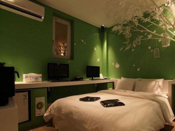 Quarto com paredes verdes, cama com roupa de cama branca, escrivaninha com um computador e janela com vista para árvores.