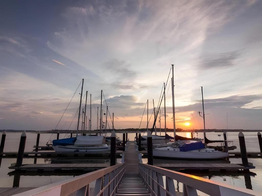 A pier leading to a marina with boats docked, a sunset in the background, and calm water.