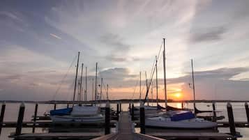 A pier leading to a marina with boats docked, a sunset in the background, and calm water.