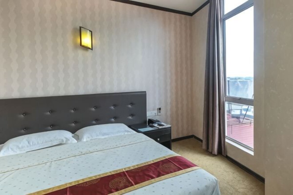 A hotel room with a large bed, a nightstand, a wall-mounted lamp, and a window with a view.
