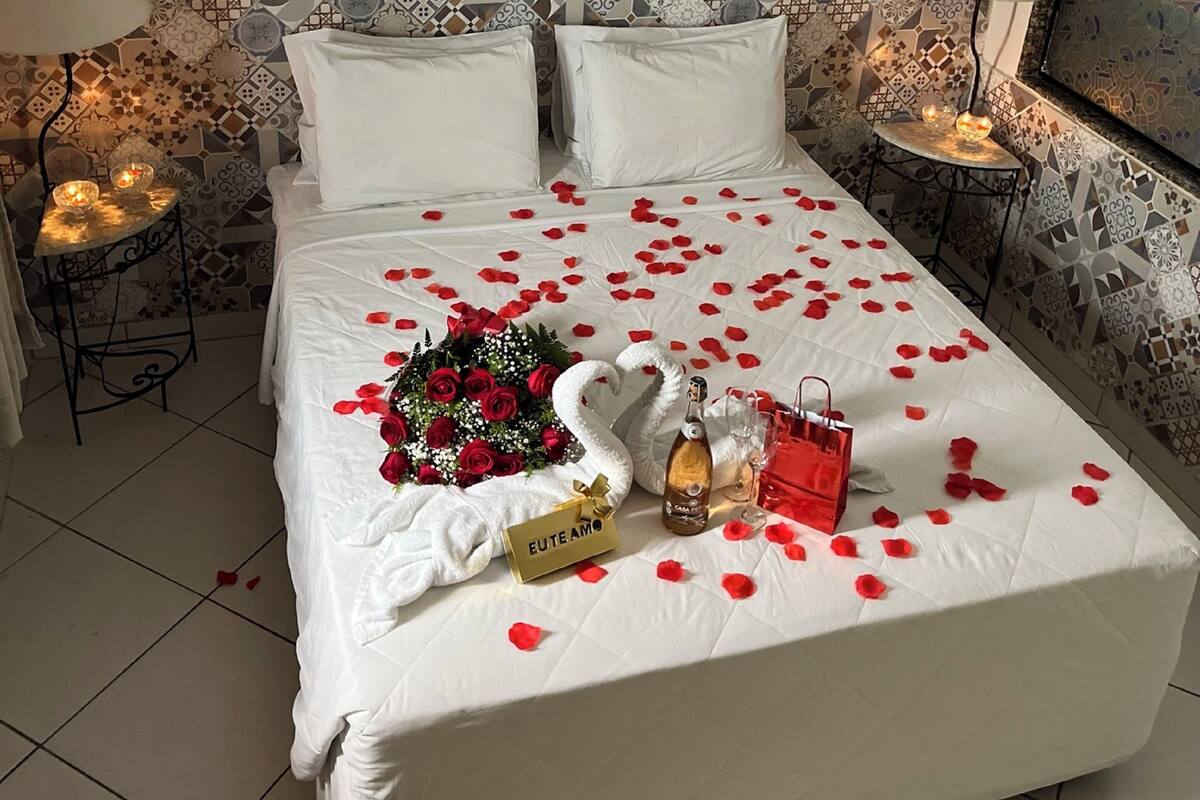 Una habitación de hotel con una cama grande, un ramo de rosas rojas, una botella de champán y una bolsa de regalo roja.