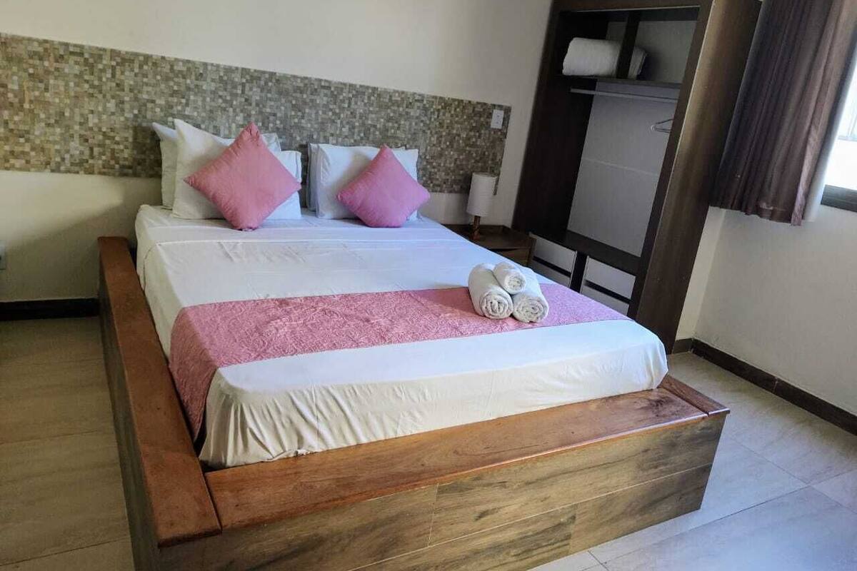 Una habitación de hotel con una cama de madera, ropa de cama blanca, almohadas rosadas y una manta rosa.
