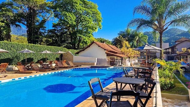 Zona de piscina con sillas de descanso y sombrillas, rodeada de exuberante vegetación y un pequeño edificio.