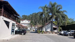 Una calle pavimentada con coches estacionados, un edificio de fachada blanca y tejado rojo, y un árbol grande al fondo.