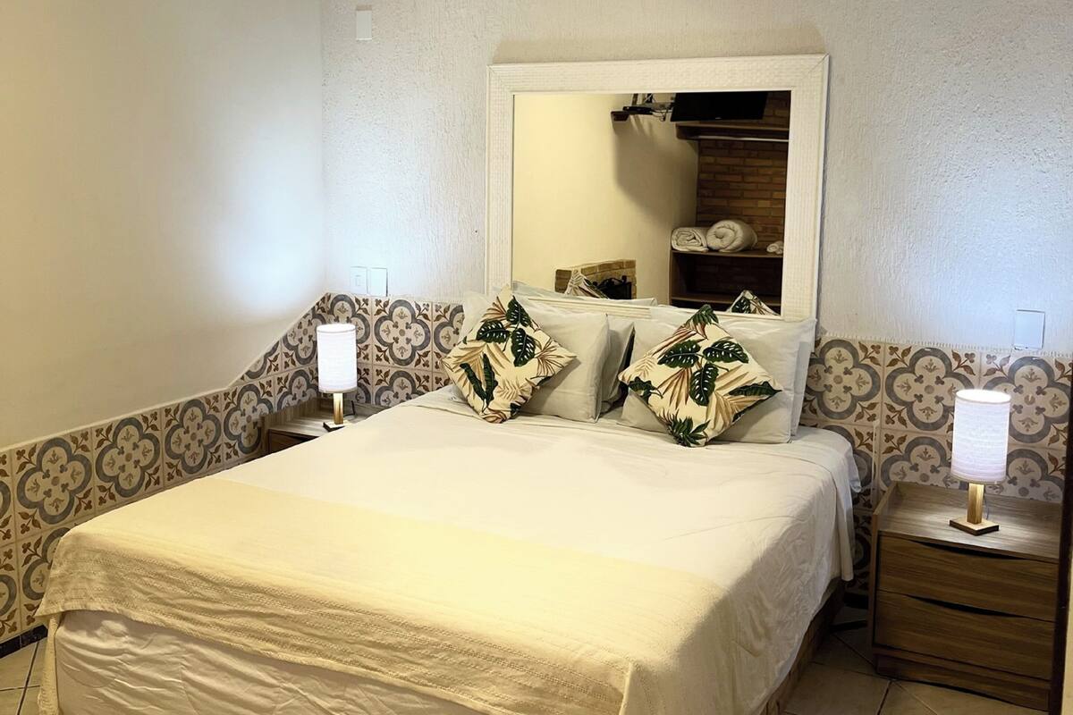 Una habitación de hotel con una cama grande, dos mesitas de noche, un espejo sobre la cama y un equipo de aire acondicionado en la pared.
