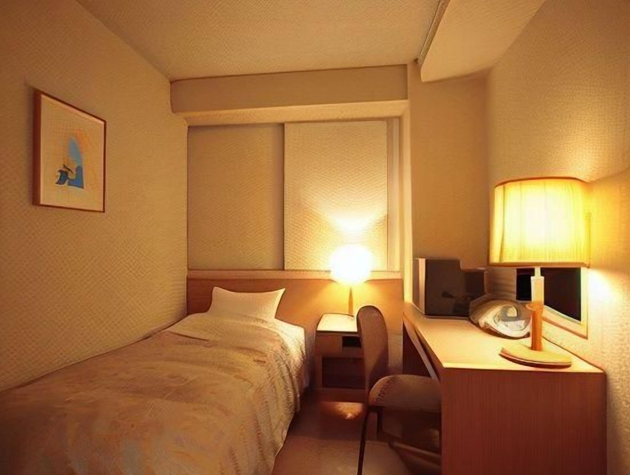 A hotel room with a bed, desk, chair, lamp, and a framed picture on the wall.