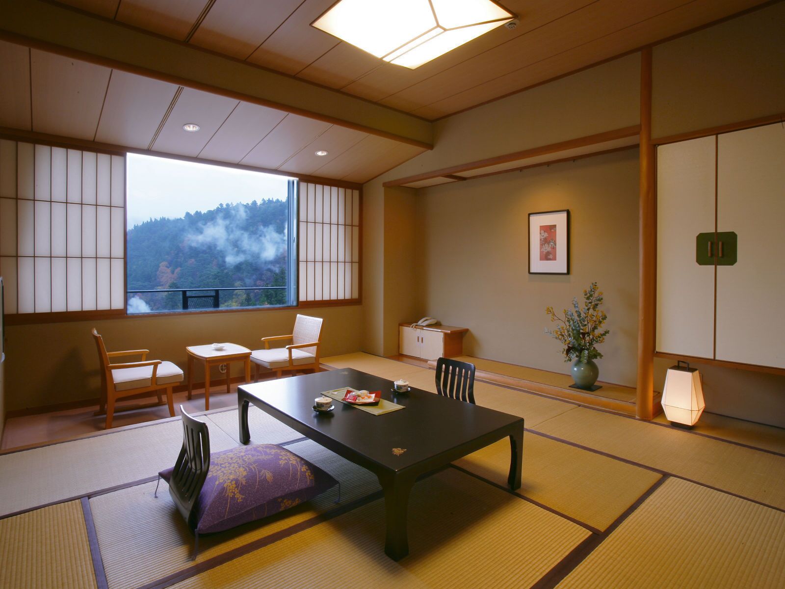 Una habitación japonesa tradicional con suelo de tatami, una mesa baja, sillas, un ventanal con vistas a un bosque y un pequeño armario con un teléfono.