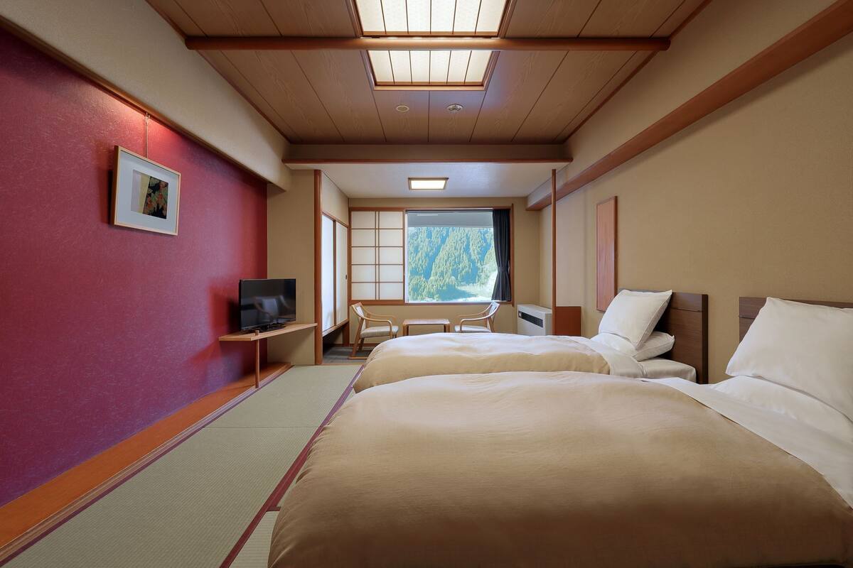 A hotel room with two beds, a TV, a desk, and a view of a forest.