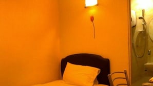 Een kleine kamer met een enkel bed, nachtkastje, stoel en een badkamer zichtbaar achter een glazen wand.