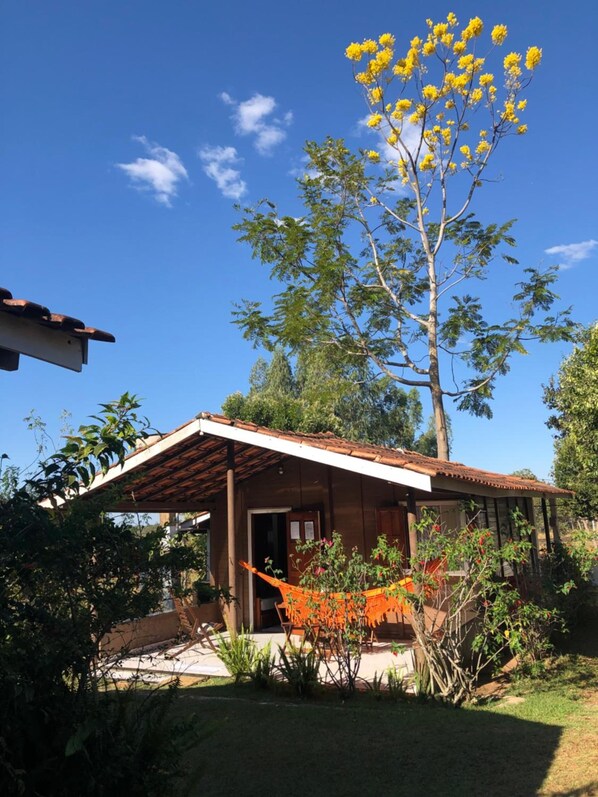 A wooden house with a tiled roof, a hammock, and a tree with yellow flowers. - Pousada Jardim da Chapada (Chapada dos Guimarães)