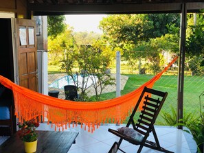 A hammock chair and a wooden bench in a covered outdoor area with a view of a swimming pool and lush greenery. - Pousada Jardim da Chapada (Chapada dos Guimarães)