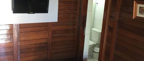 A compact room with a bed, a mounted TV, a wooden wall, and a visible bathroom.