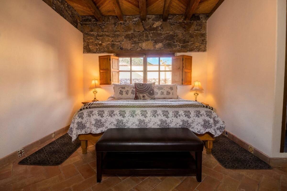 Een slaapkamer met een stenen plafond, een bed met een bloemenbedekking, twee nachtlampjes en een bankje.