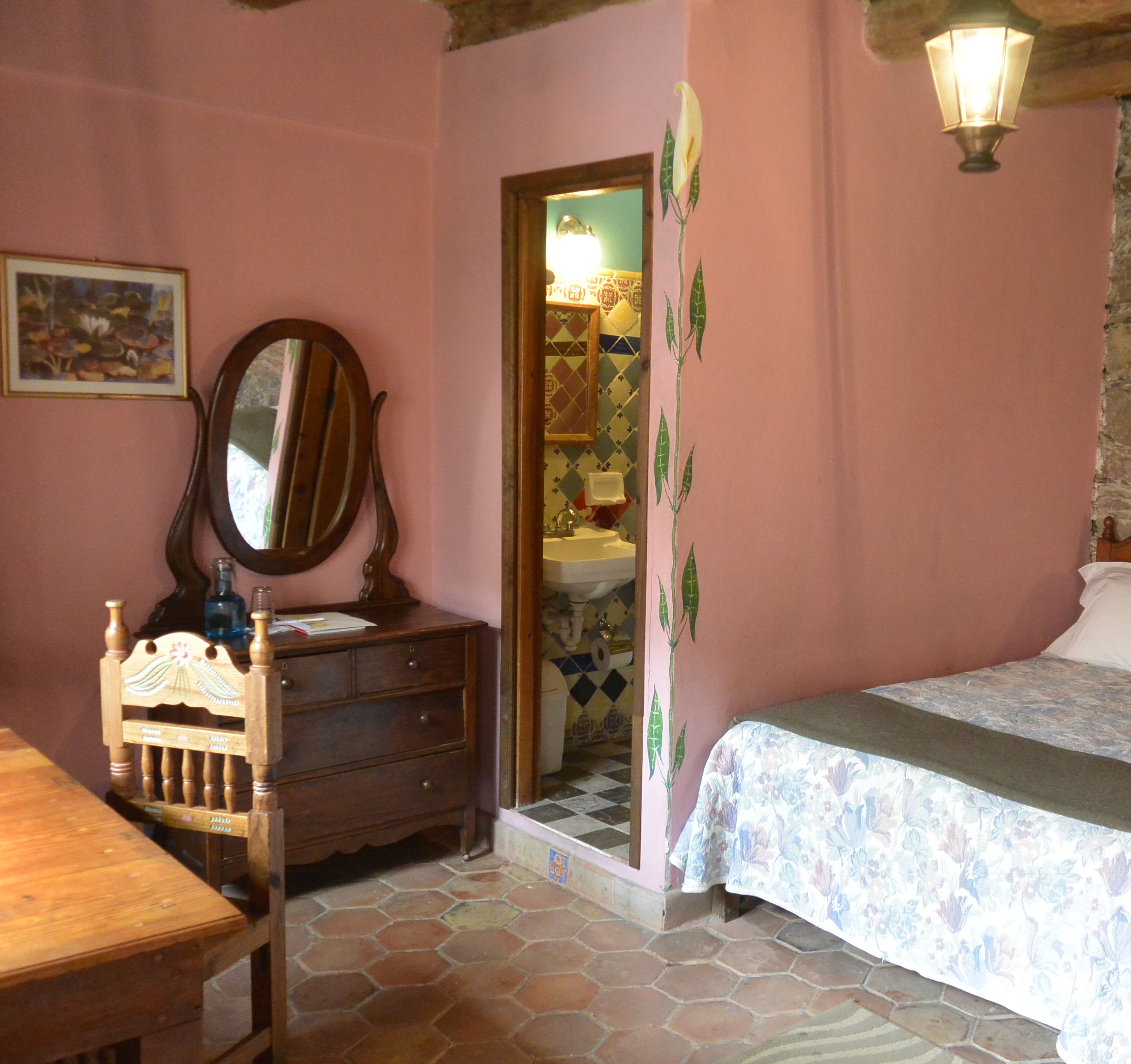 Une chambre avec une commode en bois, une chaise, un lit recouvert d’une couvre-lit à motifs floraux, un miroir et une salle de bain visible à travers une porte ouverte.