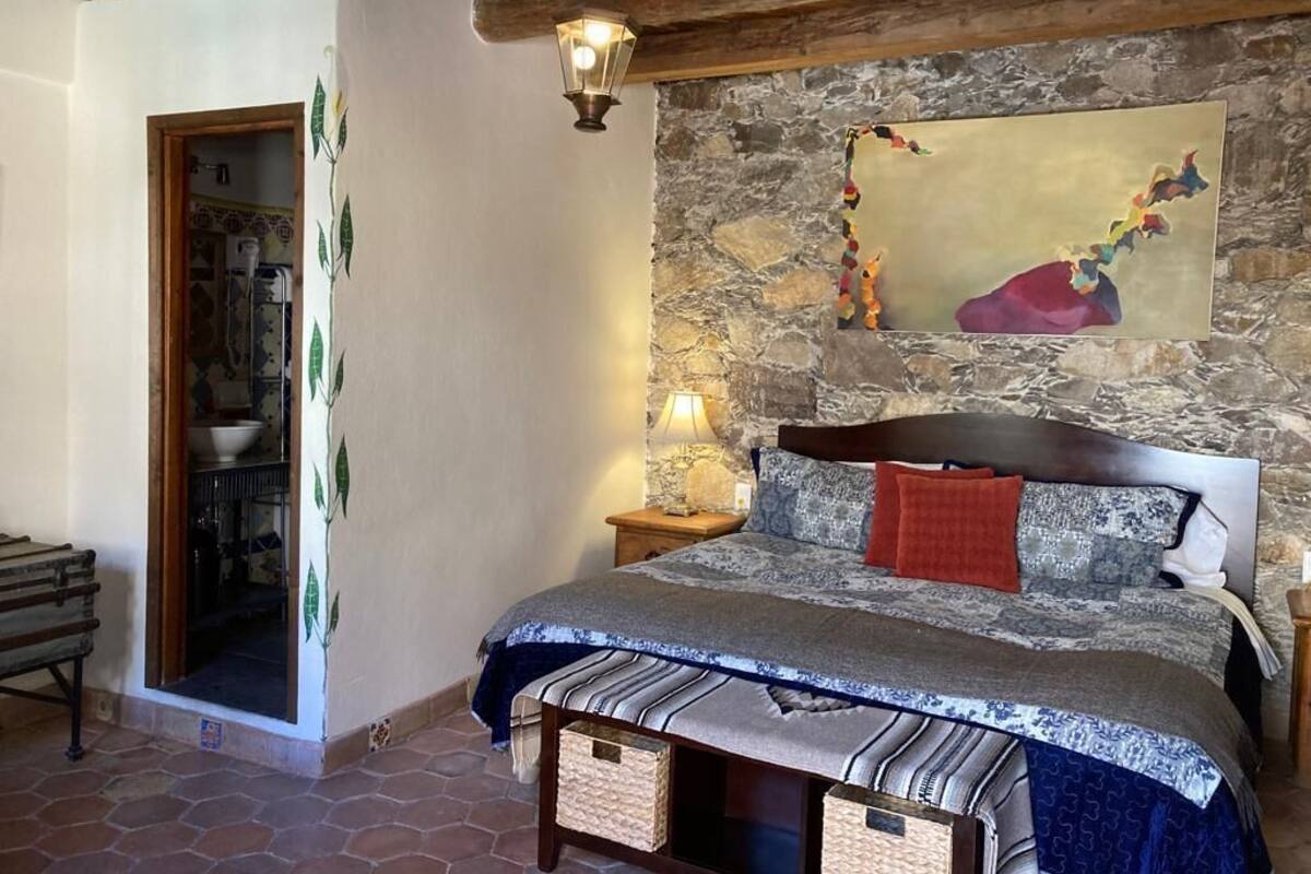 Een slaapkamer met een stenen muur, een bed met beddengoed met een patroon, een nachtkastje met een lamp en een houten bankje.