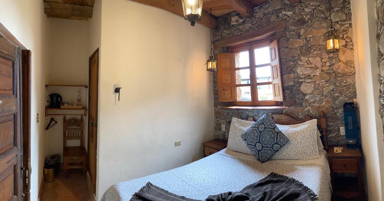 Quarto com parede de pedra, cama de madeira, janela com vista e mesa de cabeceira com abajur.