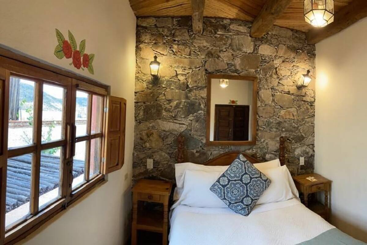 Een slaapkamer met een stenen muur, een bed met opvallende kussens, een houten nachtkastje, een spiegel en een raam met houten luiken.