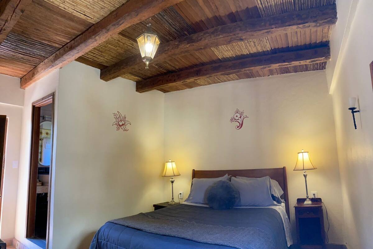 Een slaapkamer met een houten plafond, een bed met een blauwe deken, twee bedlampen en bloemmotieven op de muur.
