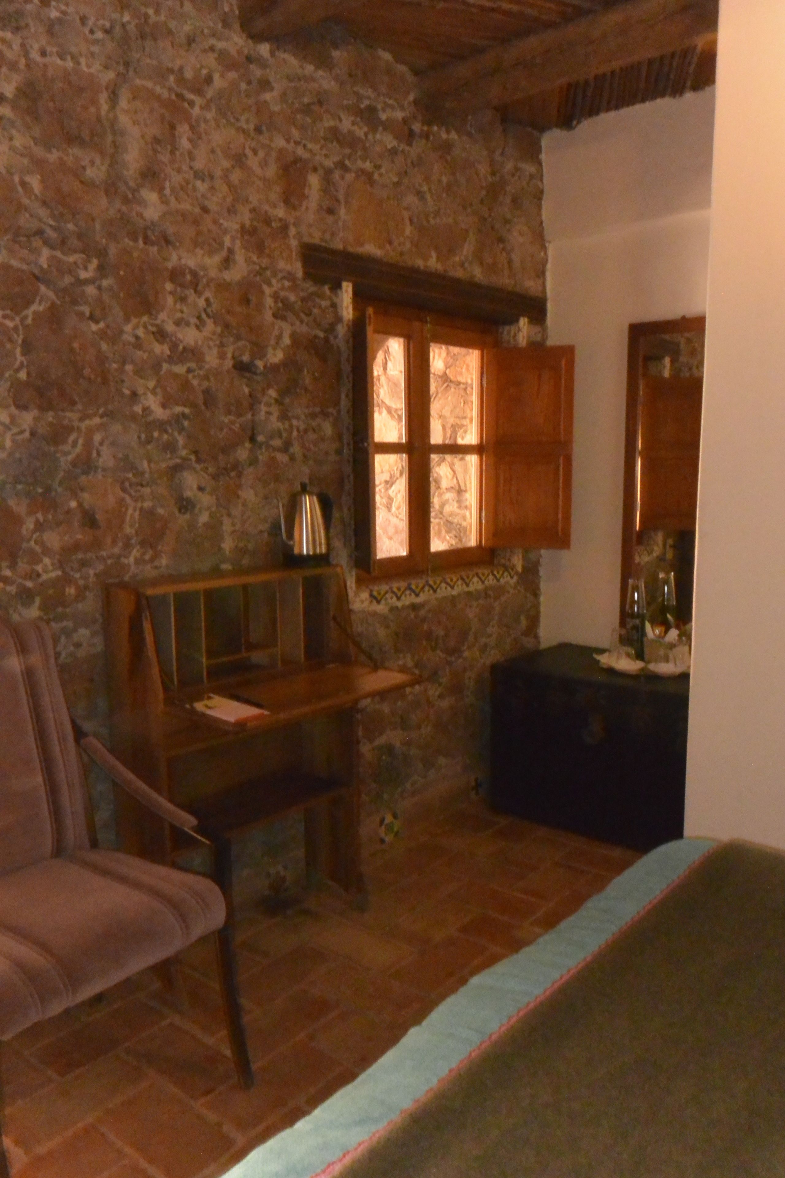 A rustic room with a stone wall, wooden furniture, a window with open shutters, and a small table with a kettle.
