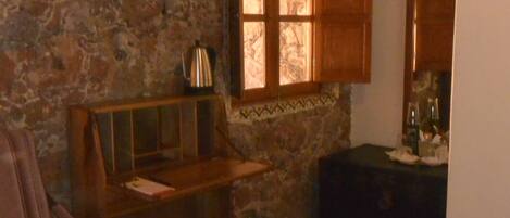 A rustic room with a stone wall, wooden furniture, a window with open shutters, and a small table with a kettle.