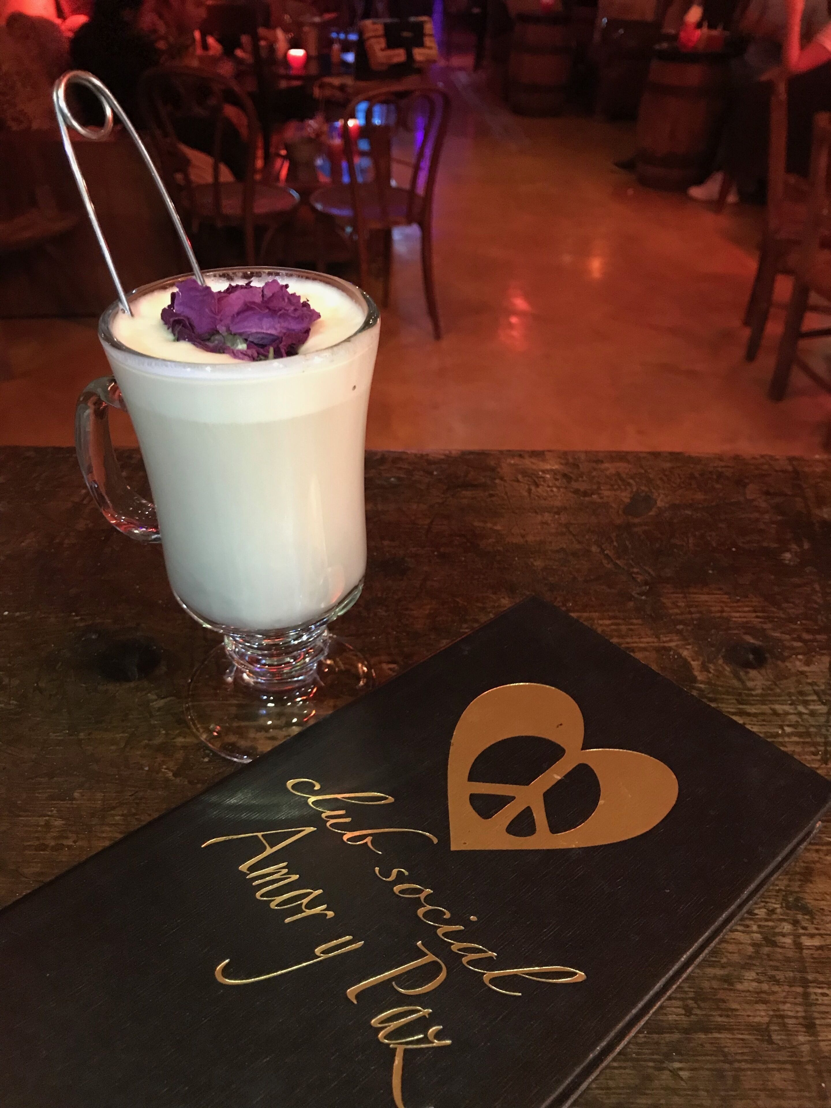 A glass of a creamy beverage with a flower garnish on a wooden table. In the background, there are chairs and tables, suggesting an indoor setting.
