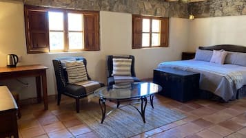 A cozy room with a stone wall, wooden ceiling beams, a bed with a blue bedspread, two upholstered chairs, a glass coffee table, and a small table with a kettle.