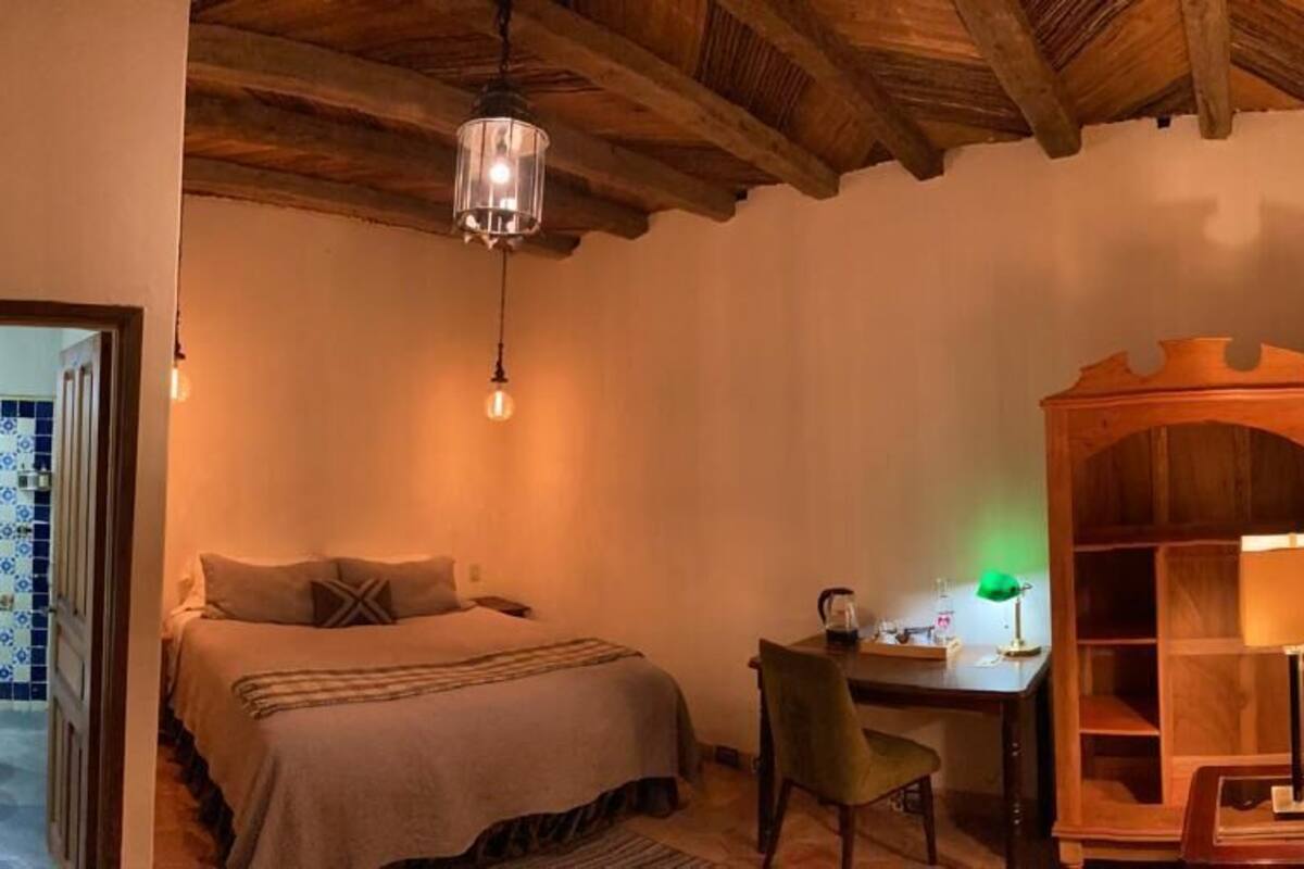 Een knus ingerichte kamer met een bed, een bureau en een stoel. Er is een houten plank en een hangende lamp.