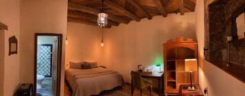 Quarto aconchegante com uma cama, uma escrivaninha e uma cadeira. Há uma estante de madeira e um lustre.