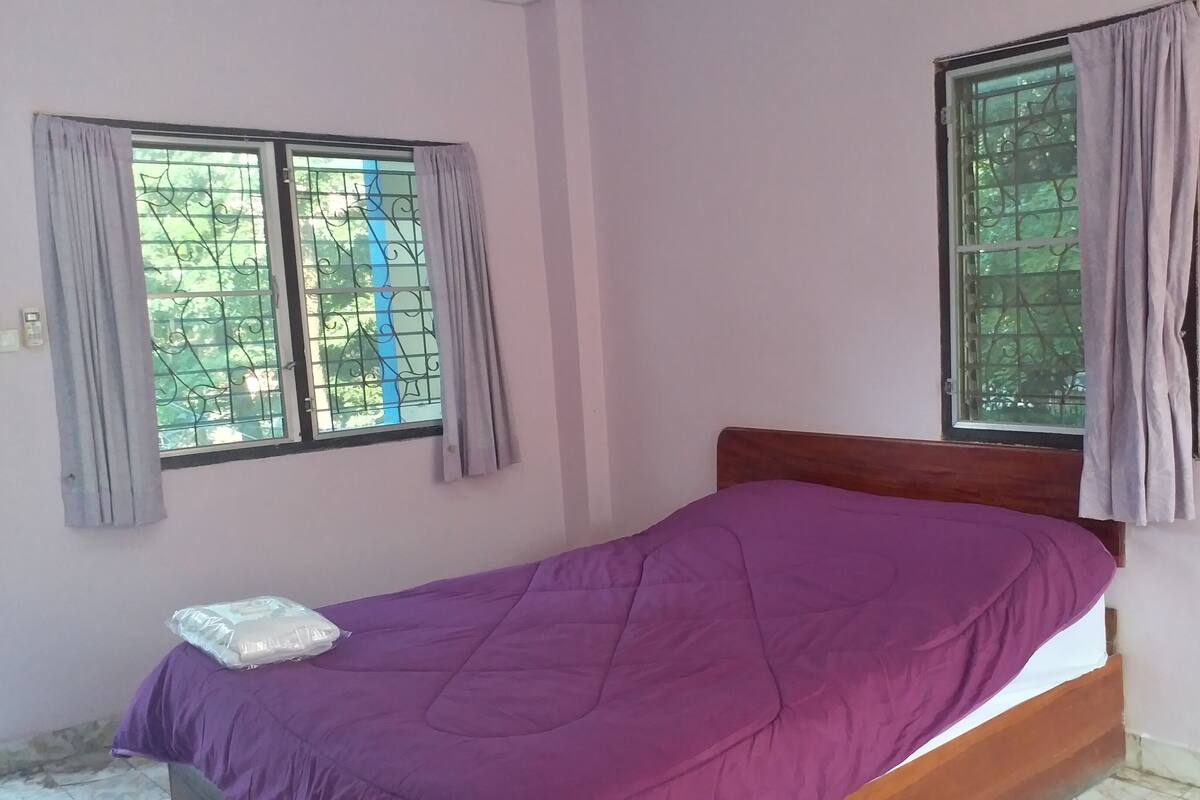 Ein kleines Zimmer mit einem Einzelbett, einer violetten Tagesdecke, zwei Fenstern mit Metallgitter und Vorhängen sowie einem Holzkopfende.