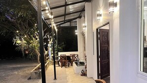 An outdoor patio area with a round table and four wooden chairs, surrounded by a metal roof with string lights. - 99 Inn Hotel (Pak Chong)