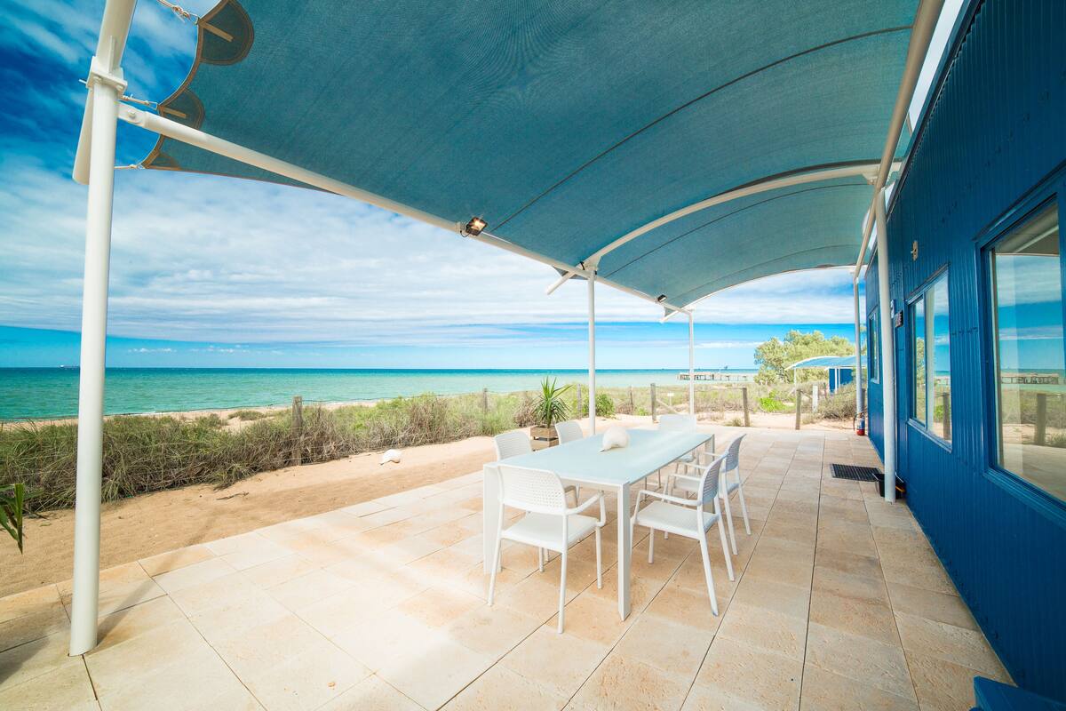 An outdoor patio with a table and chairs, a beach view, and a blue building.