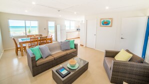 Un vaste espace de vie avec une grande fenêtre donnant sur la plage, un coin repas avec des meubles en bois et une petite cuisine.