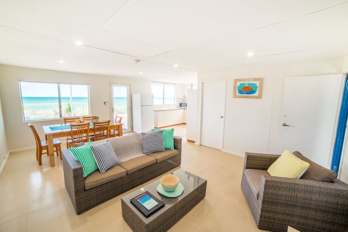 A spacious living area with a large window overlooking the beach, a dining area with wooden furniture, and a kitchenette.