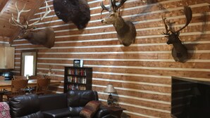Interior de una cabaña de troncos con un sofá de cuero, ciervos montados como trofeo, una estantería y un televisor.