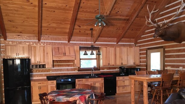Interior rústico de una cabaña con paredes de madera, una zona de comedor con una mesa cubierta con colcha y un ventilador de techo.