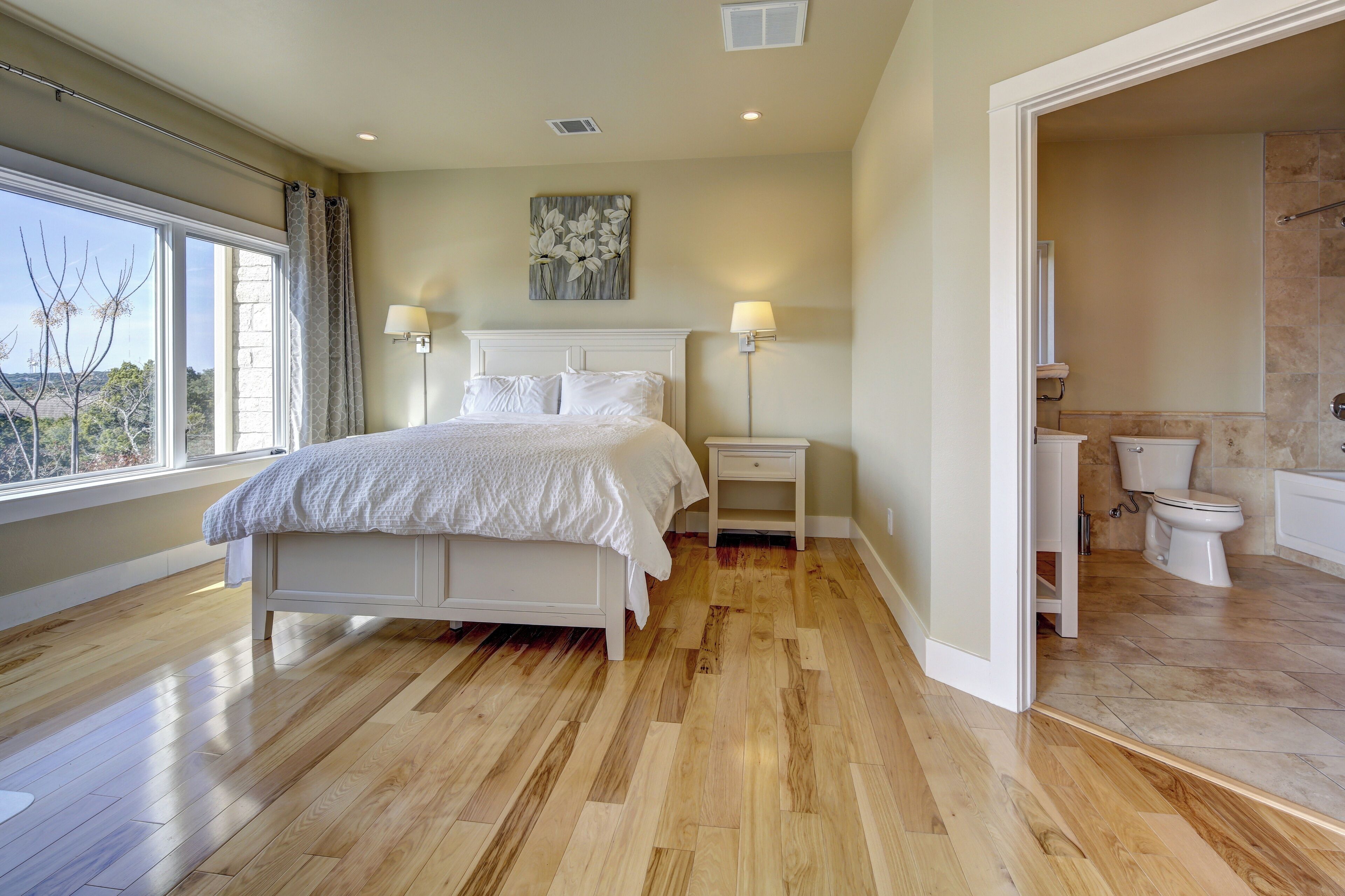 A bedroom with a large bed, two bedside tables, a nightstand, a window with curtains, and a view of the outdoors.