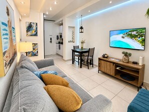 A modern living room with a sofa, a dining table with chairs, a TV mounted on the wall, and a kitchen in the background.