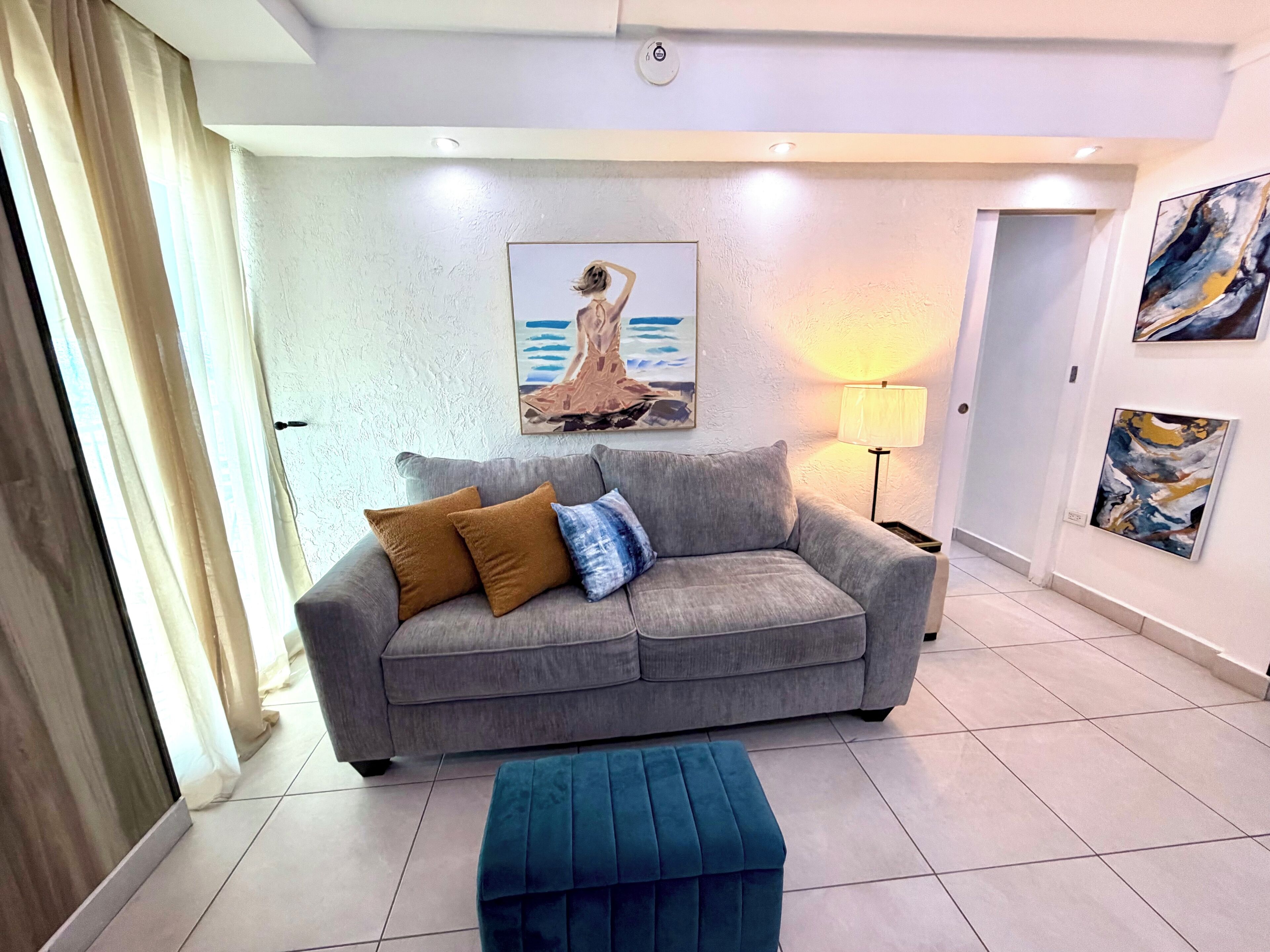 A modern living room with a grey sofa, two cushions, a blue ottoman, and a painting on the wall.