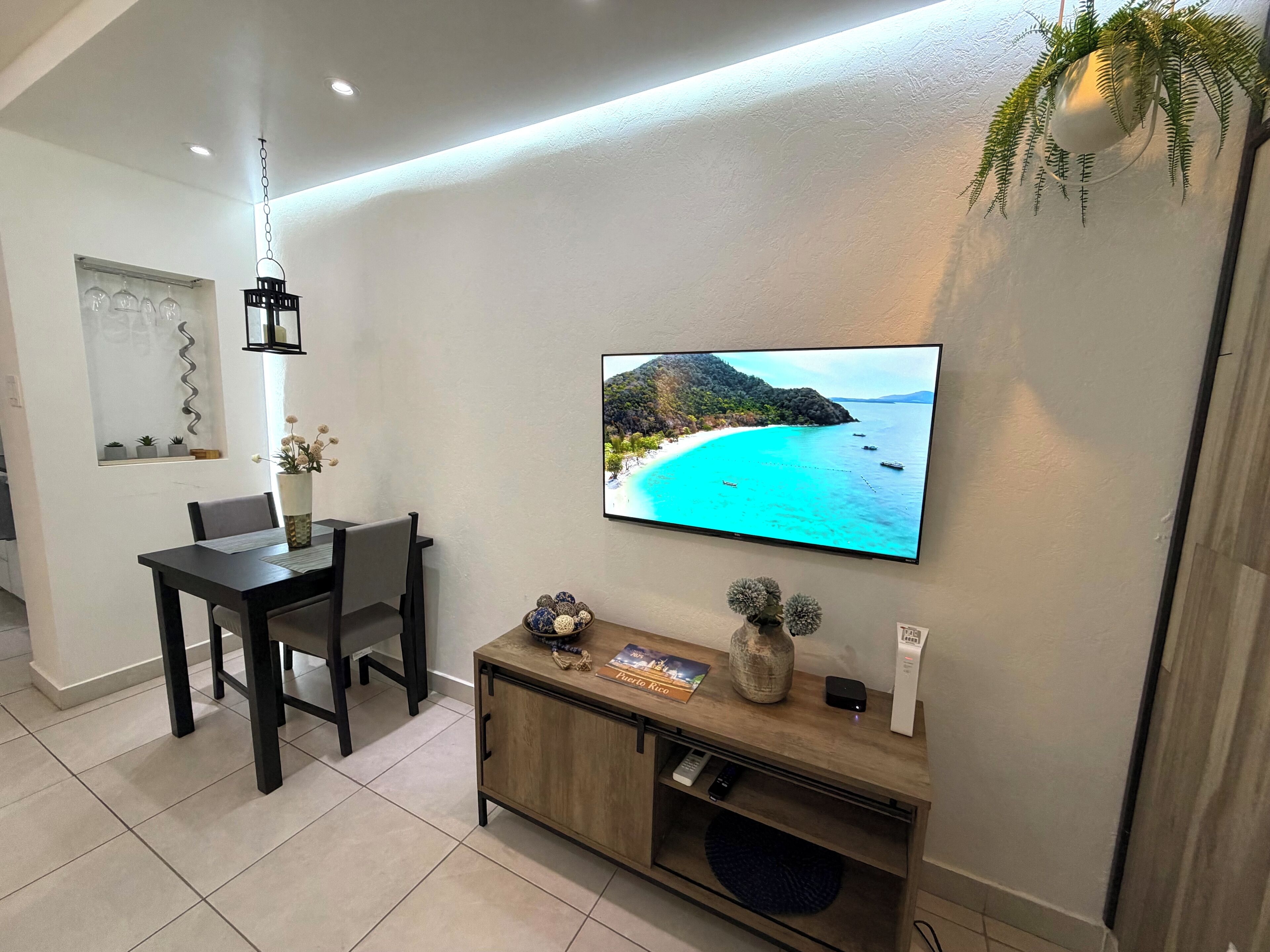 Una sala moderna con un televisor de pantalla plana mostrando una escena de playa, un comedor con mesa y sillas, y un mueble pequeño con objetos decorativos.