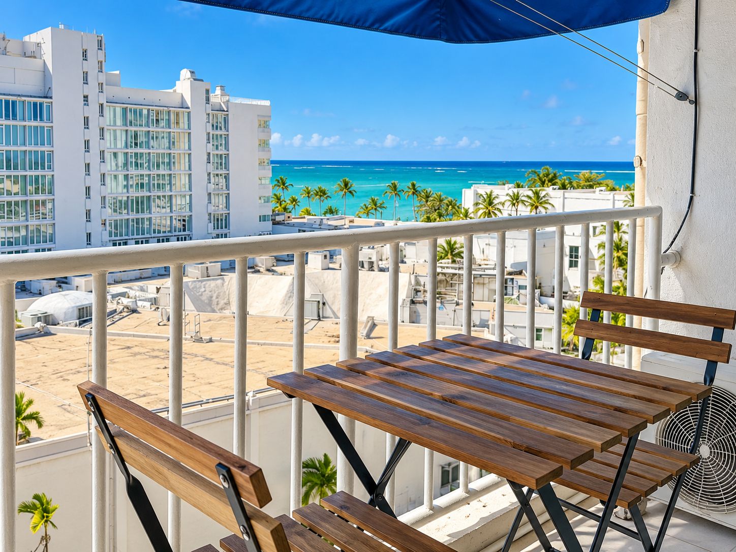A balcony with a wooden table and chairs, overlooking a beach and ocean with buildings in the background.
