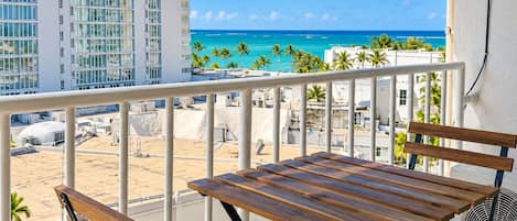 A balcony with a wooden table and chairs, overlooking a beach and ocean with buildings in the background.