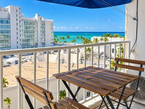 A balcony with a wooden table and chairs, overlooking a beach and ocean with buildings in the background.