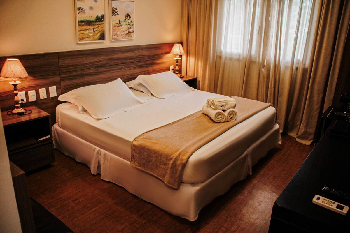 A hotel room with a neatly made bed, two bedside lamps, a headboard with artwork, and a television on the right.