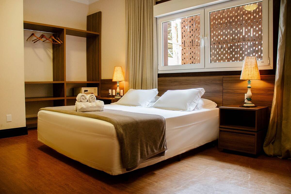 A neatly made bed with a brown blanket, two bedside lamps, a wooden nightstand, a wardrobe, and a window with a view of a wooden fence.