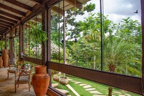 A room with large windows, wooden furniture, and a view of a lush garden. - Pousada Casa Da Quineira (Chapada dos Guimarães)